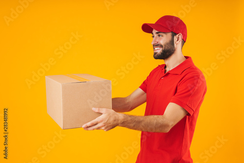 Image of pleased delivery man in red uniform smiling and holding packaging box