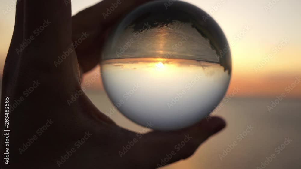 Male hand holds a glass ball on a background of sunset at sea. Dramatic cinematic shot. Silhouette. Male hand holds a glass ball on a background of sunset at sea. Dramatic cinematic shot. Silhouette.