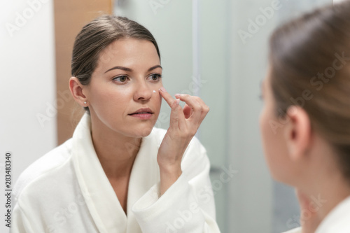 Woman standing in bathroom, applying cream on face