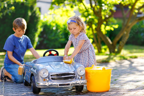 Two happy children washing big old toy car in summer garden, outdoors. Brother boy and little sister toddler girl cleaning car with soap and water, having fun with splashing and playing with sponge. photo