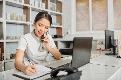 Smiling young Vietnamese female receptionist answering phone call and checking schedule in planner