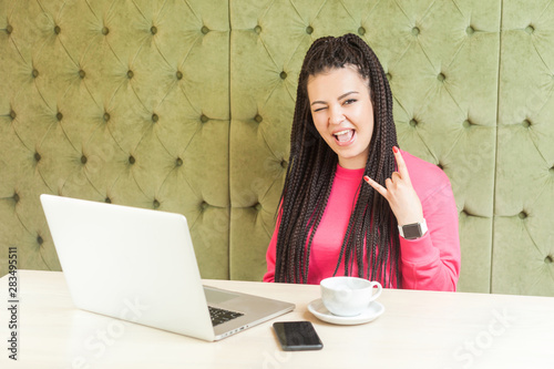 Portrait of attractive young girl freelancer with black dreadlocks hairstyle in pink blouse sitting in cafe, working on laptop winking and showing rock sign and screaming, looking at camera. indoor photo