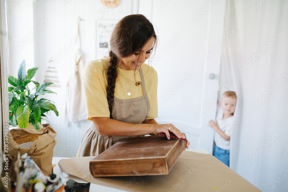 woman with braid sanding a wooden box while her son peeks from behind the door woman with braid sanding a wooden box while her son peeks from behind the door