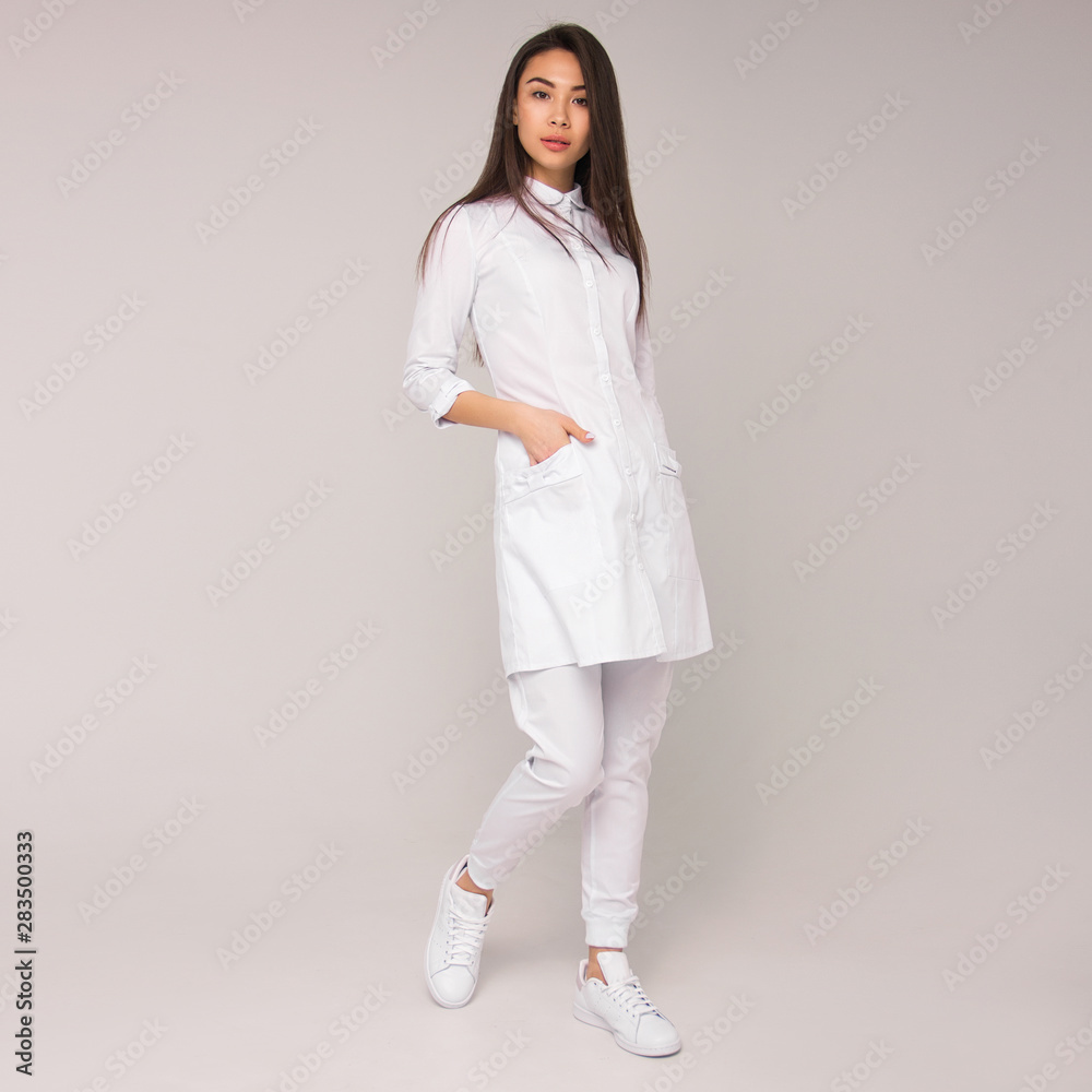 young stylish happy nurse with long brunette hair is standing with hand in her pocket, looking straight and smiling on white wall background. medical fashion concept. free space young stylish happy nurse with long brunette hair is standing with hand in her pocket, looking straight and smiling on white wall background. medical fashion concept. free space