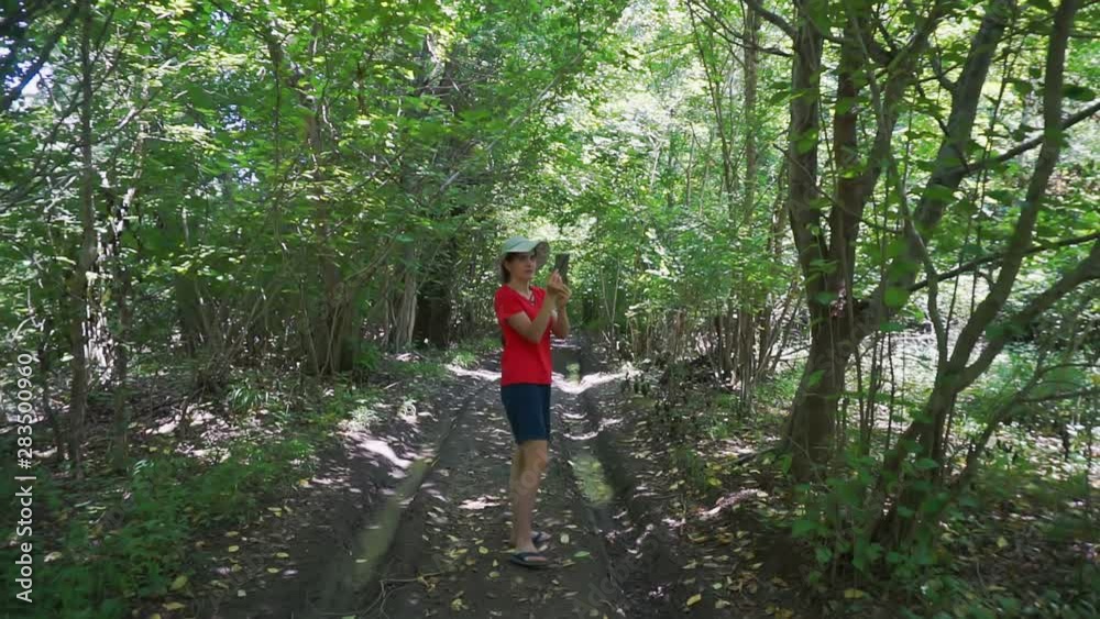 young woman on a walk in a forest grove on a sunny summer day.girl takes pictures on a modern gadget.tourist takes pictures on an electronic device young woman on a walk in a forest grove on a sunny summer day.girl takes pictures on a modern gadget.tourist takes pictures on an electronic device