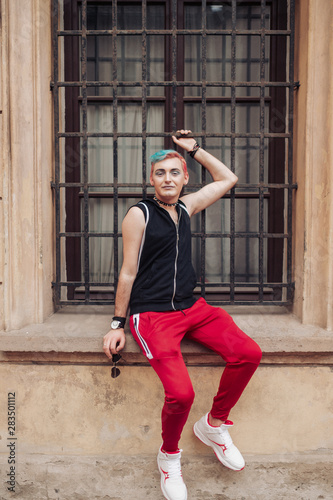 Gay man in stylish clothing with colorful hairstyle posing with passion on window with metal grid.