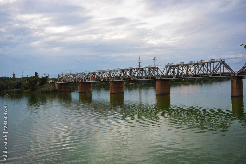 The gray Samara railway bridge over the surface of the Samara river with a beautiful landscape, green trees, bushes and greenery. Pictures of the river landscape with an unusual sky and bright clouds. The gray Samara railway bridge over the surface of the Samara river with a beautiful landscape, green trees, bushes and greenery. Pictures of the river landscape with an unusual sky and bright clouds.