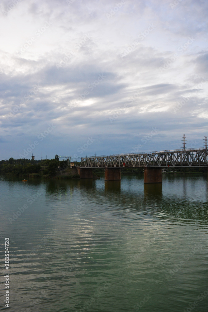 The gray Samara railway bridge over the surface of the Samara river with a beautiful landscape, green trees, bushes and greenery. Pictures of the river landscape with an unusual sky and bright clouds.