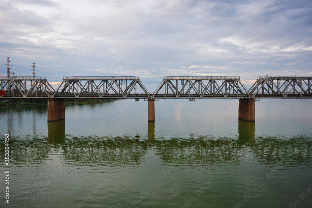 The gray Samara railway bridge over the surface of the Samara river with a beautiful landscape, green trees, bushes and greenery. Pictures of the river landscape with an unusual sky and bright clouds. The gray Samara railway bridge over the surface of the Samara river with a beautiful landscape, green trees, bushes and greenery. Pictures of the river landscape with an unusual sky and bright clouds.