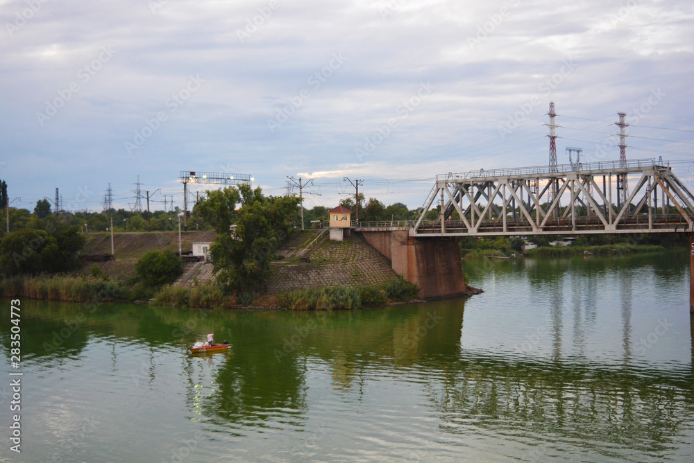 The gray Samara railway bridge over the surface of the Samara river with a beautiful landscape, green trees, bushes and greenery. Pictures of the river landscape with an unusual sky and bright clouds. The gray Samara railway bridge over the surface of the Samara river with a beautiful landscape, green trees, bushes and greenery. Pictures of the river landscape with an unusual sky and bright clouds.