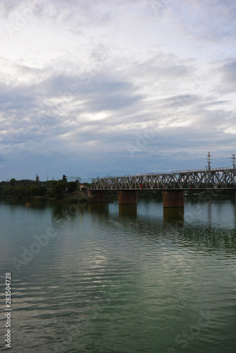 The gray Samara railway bridge over the surface of the Samara river with a beautiful landscape, green trees, bushes and greenery. Pictures of the river landscape with an unusual sky and bright clouds. © Daria Katiukha