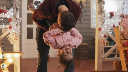 Happy father having fun with her kid daughter at Christmas porch. Dad tickling her little daughter in slow motion. Father and daughter in pink pullover at Christmas day. Close-up shot