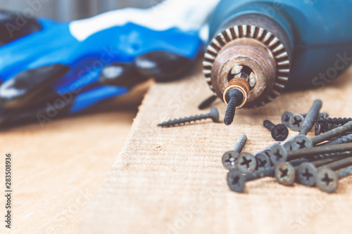 A steel screw is placed on an electric drill near scattered screws on a wooden board background. The concept of tools and repair work. Steel screws.