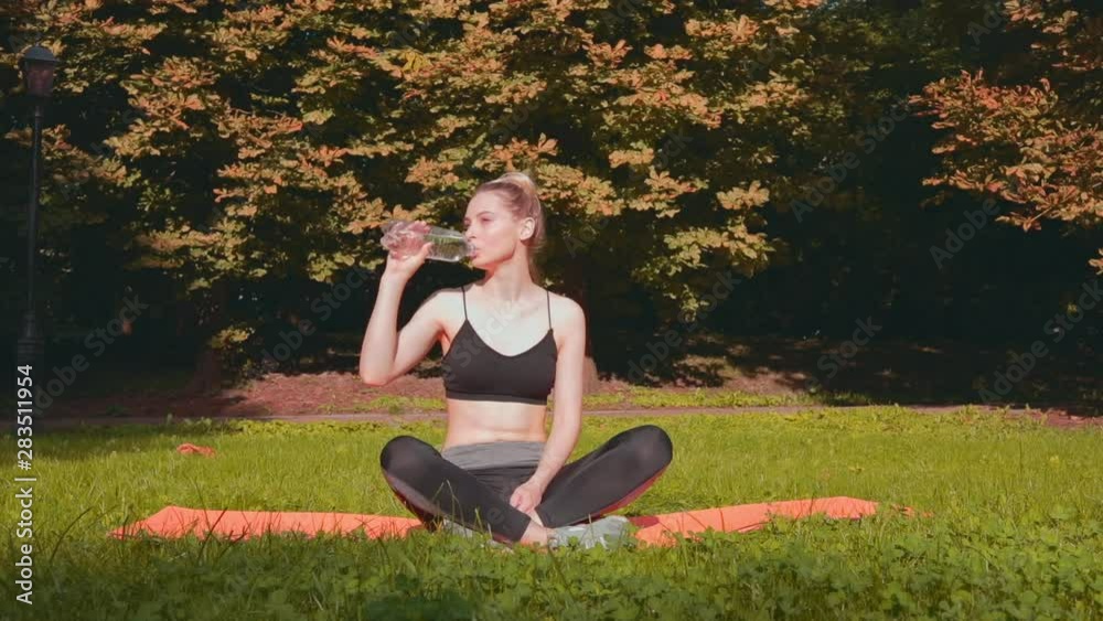 Young sporty girl drink water in morning sunny park. Woman doing yoga in the park. Athlete woman doing stretching exercise in summer park. Healthy sports lifestyle. Young sporty girl drink water in morning sunny park. Woman doing yoga in the park. Athlete woman doing stretching exercise in summer park. Healthy sports lifestyle.