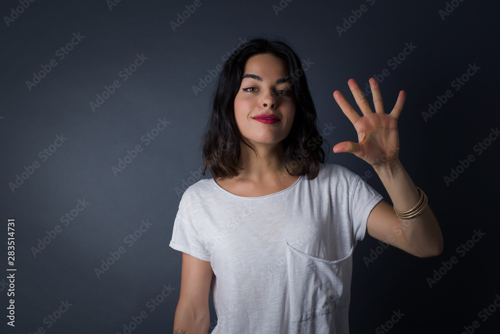 Young woman standing against gray wall showing and pointing up with fingers number five while smiling confident and happy.