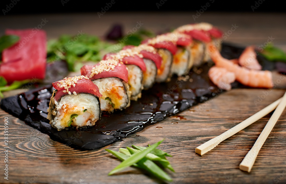 Sushi covered with tuna and sesame seeds on sloppy lubricate stone plate. Focus on details. Front view. Bamboo chopsticks, green onion on table. Tuna piece, shrimps, salad leaves on blurred background Sushi covered with tuna and sesame seeds on sloppy lubricate stone plate. Focus on details. Front view. Bamboo chopsticks, green onion on table. Tuna piece, shrimps, salad leaves on blurred background