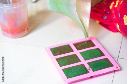 Pouring green colored soap mix with red swirls from a vessle with snout into a pink mold for making final home made hand made soap bars photo