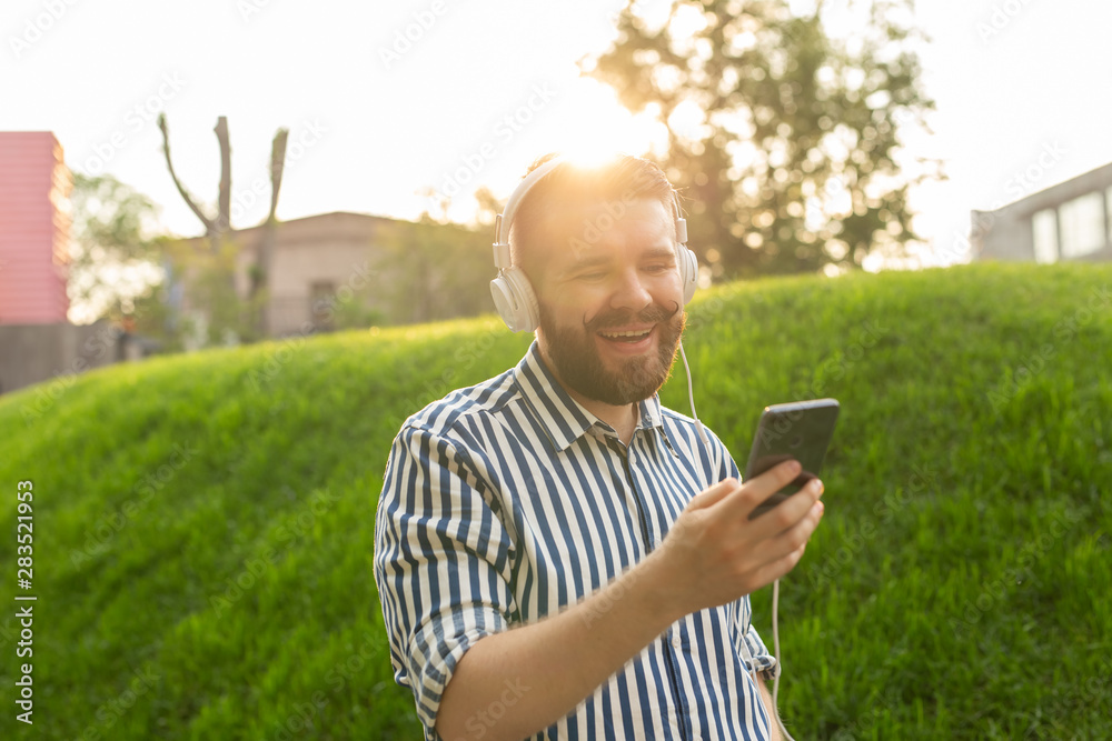 Portrait of a positive young man with a mustache and beard listening to an audiobook while walking through the park. Leisure learning concept. Portrait of a positive young man with a mustache and beard listening to an audiobook while walking through the park. Leisure learning concept.
