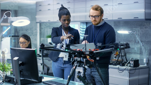 Caucasian Male and Black Female Engineers Working on a Drone Project with Help of Laptop and Taking Notes. He Works in a Bright Modern High-Tech Laboratory.