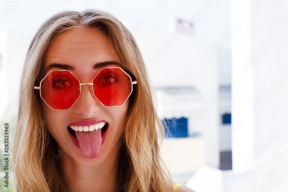 Portrait of beautiful emotional young woman in summer red sunglasses on a sea background shows tongue Portrait of beautiful emotional young woman in summer red sunglasses on a sea background shows tongue