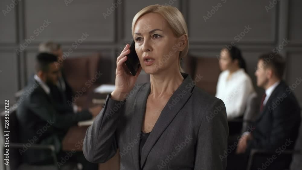 Chest-up shot of middle-aged Caucasian businesswoman in formal suit standing in meeting room and talking on phone with CEO and nodding head, and executive colleagues sitting and talking in background Chest-up shot of middle-aged Caucasian businesswoman in formal suit standing in meeting room and talking on phone with CEO and nodding head, and executive colleagues sitting and talking in background