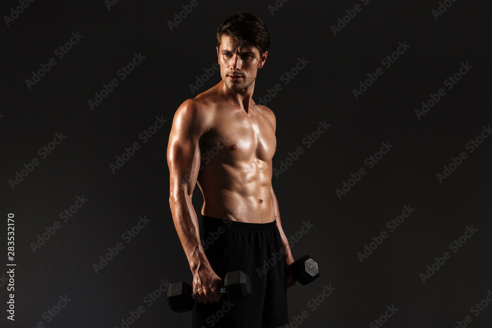 Concentrated handsome young strong sportsman posing isolated over black wall background holding dumbbells.