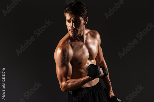 Concentrated handsome young strong sportsman posing isolated over black wall background holding dumbbells. © Drobot Dean