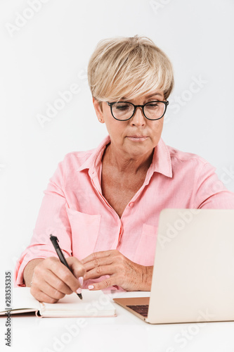 Portrait of mature middle-aged woman with short blond hair wearing eyeglasses using laptop computer and notebook