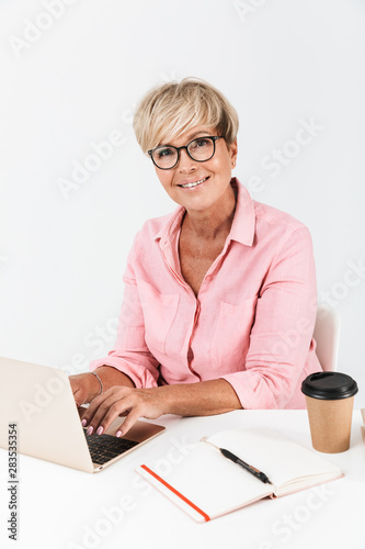 Portrait of happy middle-aged woman with short blond hair wearing eyeglasses using laptop computer at table