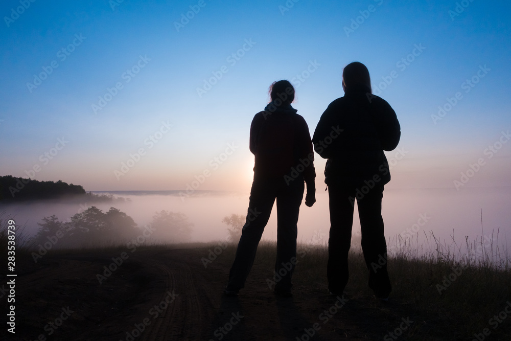 silhouette of two peple looking at sunrise on foggy valley