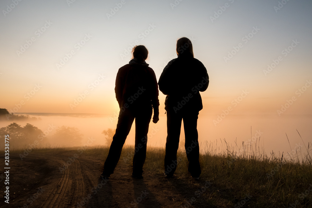 silhouette of two peple looking at sunrise on foggy valley
