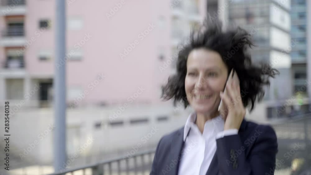 Smiling businesswoman talking on phone while walking on street. Energetic young lady wearing suit having pleasant conversation through smartphone. Communication concept Smiling businesswoman talking on phone while walking on street. Energetic young lady wearing suit having pleasant conversation through smartphone. Communication concept