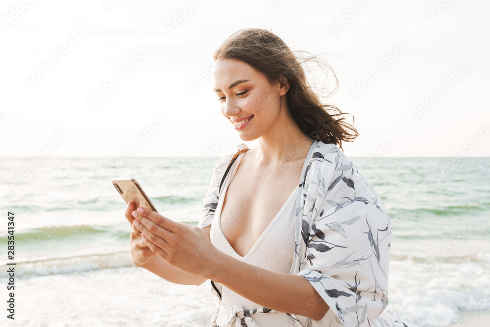 Smiling pleased happy young beautiful woman at the beach walking in a beautiful sunny morning using mobile phone.