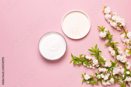 Beauty Spa concept. Opened plastic container with cream and spring White flowers on pink background Flat lay top view. Herbal dermatology cosmetic hygienic cream, organic cosmetic Natural