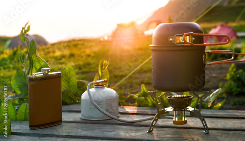 Cooking pot on camping gas stove and hip flask at sunlight background. Travel equipment at beautiful sunset