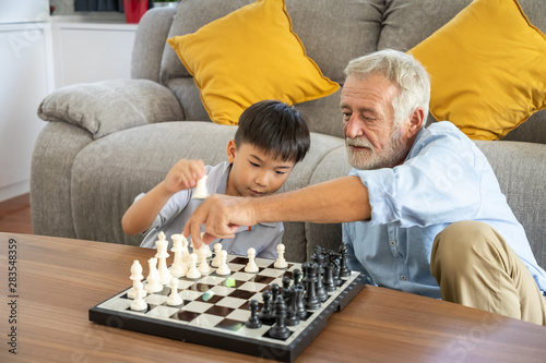 Happy boy grandson playing chess with old senior man grandfather at home