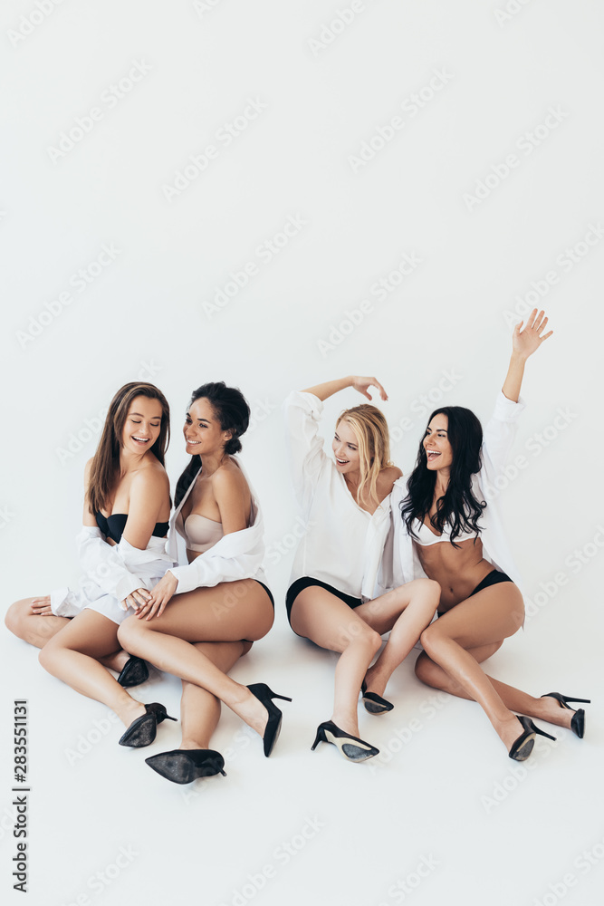 Obraz premium four smiling sexy multiethnic girls wearing white shirts and heels sitting on grey