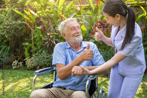 Happy nurse holding elderly man hand on wheelchair in garden at nursing home thumb up
