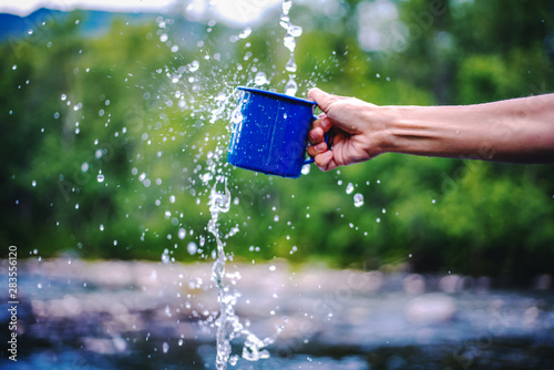Blue camping mug with clear water, splashes on a forest background. Clear water from a mountain river, ecology concept