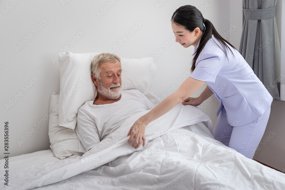 Happy nurse cover elderly man with blanket in bedroom at nursing home