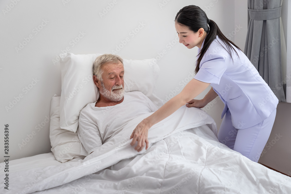 Fototapeta premium Happy nurse cover elderly man with blanket in bedroom at nursing home