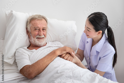Happy nurse holding elderly man hands with blanket in bedroom at nursing home looking camera