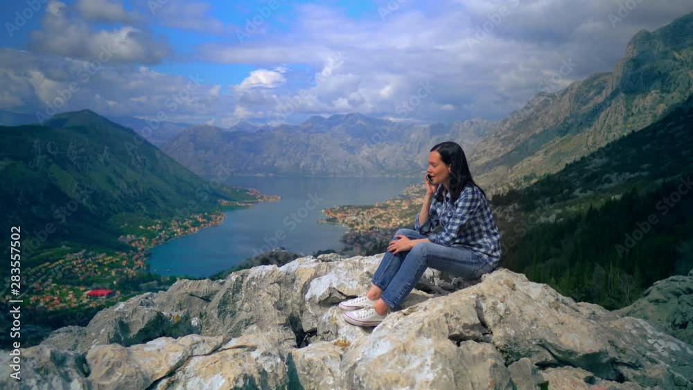 woman traveller sitting on the rock talking by phone view of the mountainous area with blue water houses in the valley surrounded trees woman traveller sitting on the rock talking by phone view of the mountainous area with blue water houses in the valley surrounded trees