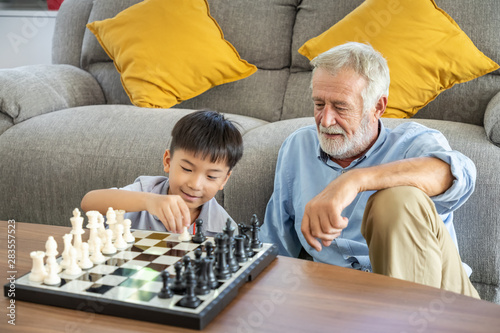 Happy boy grandson playing chess with old senior man grandfather at home