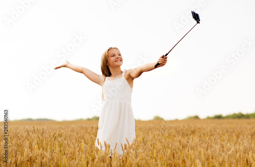 technology, summer and people concept - happy young girl in white dress taking picture by smartphone on selfie stick on cereal field