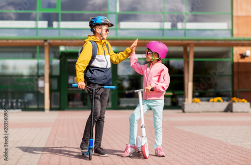 education, childhood and people concept - happy school children in helmets with backpacks riding scooters and making high five outdoors