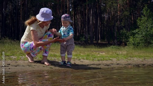Mom and her little son play near the water on the shore of a forest lake on a summer sunny day. Mom and her baby throw stones in the water and talk