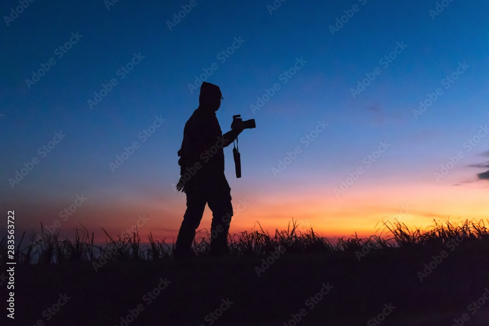 Silhouette of man taking pictures with his camera while sunset hour; against twilight sky over a mountain. Traveling and vacation concept.