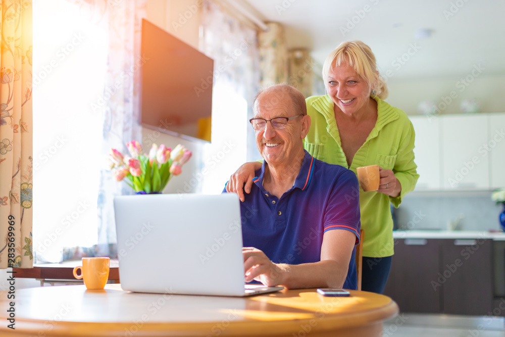 Senior couple looking at the lap top screen smiling and drinking coffee - elderly people video calling or talking by web camera - working at home, freelancing and having hobbies together concept