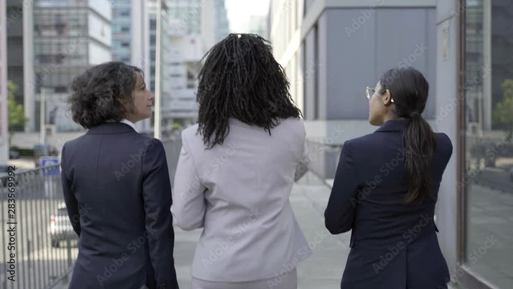 Back view of businesswomen walking on street. Rear view of professional multiethnic young female colleagues holding folders and talking while walking outside modern office building. Business concept Back view of businesswomen walking on street. Rear view of professional multiethnic young female colleagues holding folders and talking while walking outside modern office building. Business concept